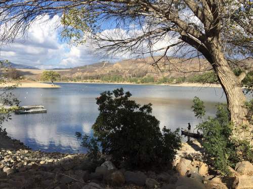 A serene lake surrounded by trees and rocky shore, with a cloudy sky and distant hills in the background.