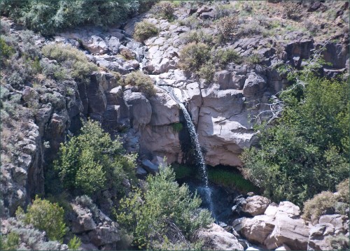 A small waterfall cascades over rocky cliffs, surrounded by lush greenery and rugged terrain.