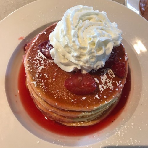 Stack of fluffy pancakes topped with whipped cream and strawberries, drizzled with strawberry sauce.