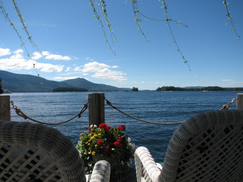 A serene lakeside view with two wicker chairs, a flower pot, and mountains in the background under a clear blue sky.