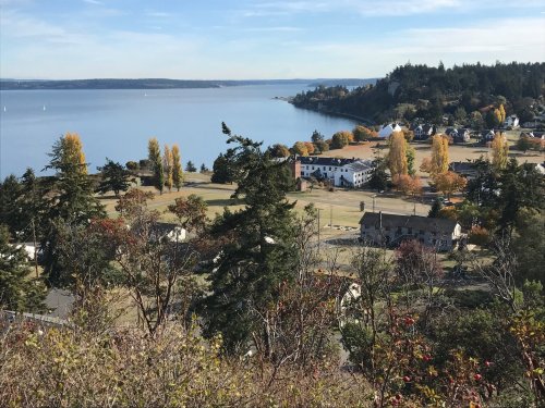 A scenic view of a calm bay surrounded by trees and autumn-colored foliage, with distant hills and buildings visible.