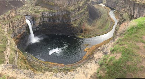 A panoramic view of a waterfall cascading into a circular pool, surrounded by steep, rocky cliffs and lush greenery.