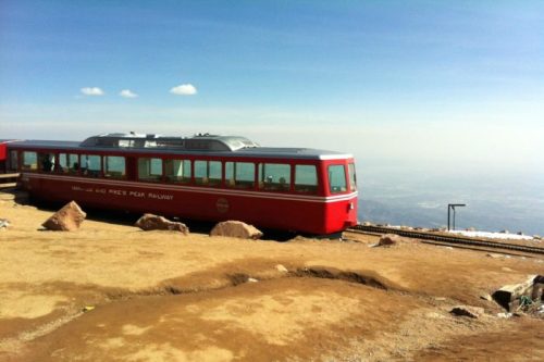A red train on a mountain track with a clear blue sky and rocky terrain in the background.