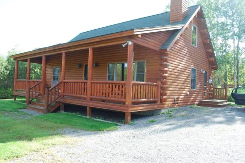 A wooden log cabin with a porch, surrounded by greenery and gravel driveway, under a clear blue sky.