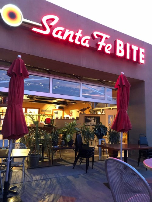 Neon sign for "Santa Fe BITE" outside a restaurant with red umbrellas and plants in the evening light.