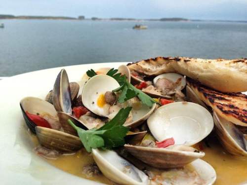 A plate of clams and seafood with herbs, set against a scenic waterfront view.