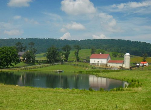 A serene farm scene with a pond, white house, red barn, silo, and green hills under a blue sky with fluffy clouds.