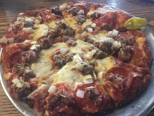 A pizza topped with pepperoni, sausage, cheese, and diced onions, served on a silver platter with a pepperoncini.