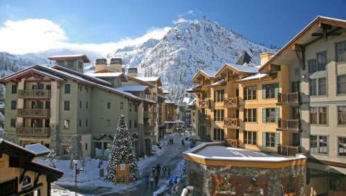 Snow-covered buildings and a decorated Christmas tree in a winter resort village, with mountains in the background.