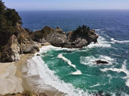 A scenic coastal view featuring turquoise waves crashing on a sandy beach surrounded by rocky cliffs and trees.
