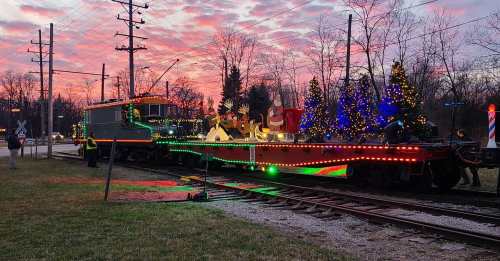 A decorated train with festive lights and holiday figures, set against a colorful sunset and trees.