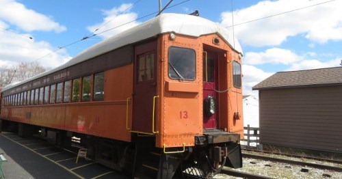 An orange and brown vintage train car parked at a station under a blue sky with clouds.