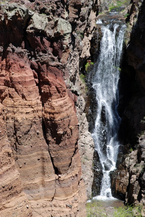 A cascading waterfall flows down rocky cliffs with colorful layers of sediment in a natural landscape.