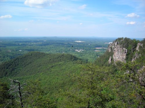 A panoramic view of lush green hills and valleys under a blue sky with scattered clouds. Rocky cliffs in the foreground.