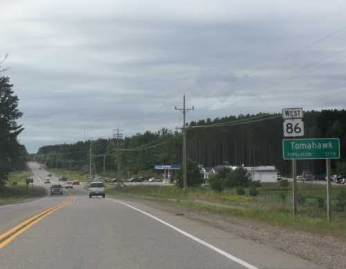 Road sign indicating the direction to Tomahawk, population 3,171, along a highway with vehicles and trees in the background.