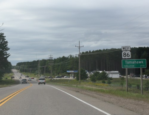 Road sign indicating the direction to Tomahawk, population 3,171, along a highway with vehicles and trees in the background.