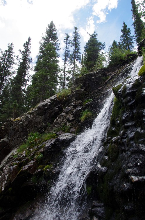 A cascading waterfall flows down rocky terrain, surrounded by lush green trees under a partly cloudy sky.
