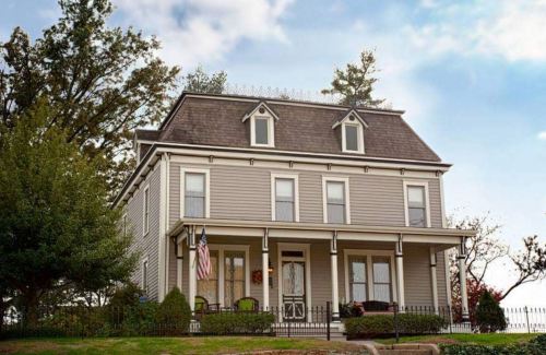 A charming two-story gray house with a sloped roof, surrounded by greenery and featuring a front porch and American flag.