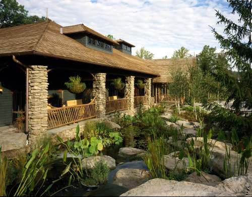 A rustic building with stone pillars, surrounded by greenery and a pond with rocks and plants.