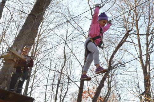 A child in a pink jacket balances on a rope bridge while an adult watches from a platform in the trees.