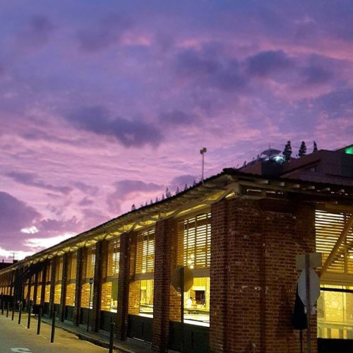 A brick building with large windows under a colorful sunset sky, casting a warm glow on the street below.