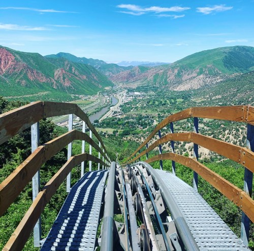 View from the top of a roller coaster track overlooking a lush valley and mountains under a clear blue sky.