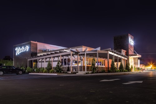 A modern restaurant exterior at night, featuring a sleek design and illuminated signage, surrounded by landscaping.