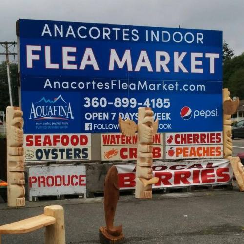 Sign for Anacortes Indoor Flea Market, featuring various food items and wooden carvings, with contact info and hours.