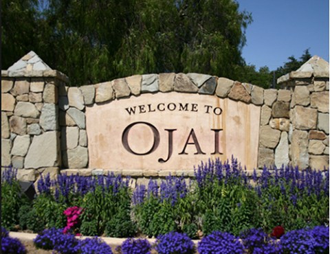 A stone sign reading "Welcome to Ojai," surrounded by vibrant purple flowers under a clear blue sky.