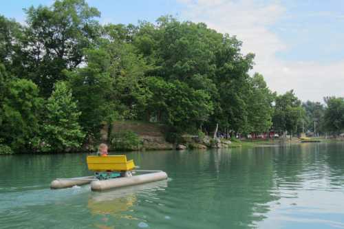 A child paddles a small yellow boat on a calm, green lake surrounded by trees.