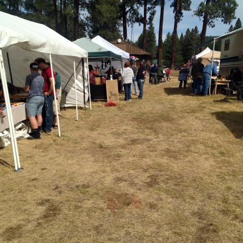 A bustling outdoor market with tents, vendors, and visitors walking along a grassy path under a clear sky.
