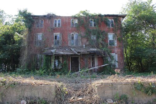 Abandoned brick building overgrown with vines and surrounded by dense vegetation, showing signs of decay.