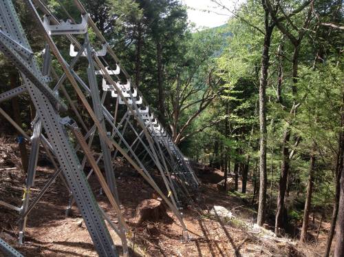 Metal structures line a forested hillside, surrounded by trees and underbrush, indicating construction or maintenance work.