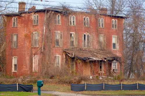 Abandoned red brick house overgrown with vines, showing signs of decay and neglect, set against a rural backdrop.