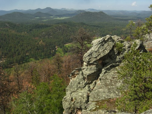 A rocky outcrop overlooks a lush green valley and distant mountains under a clear blue sky.