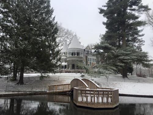 A snow-covered mansion with a turret, surrounded by trees, next to a pond with a wooden bridge in the foreground.