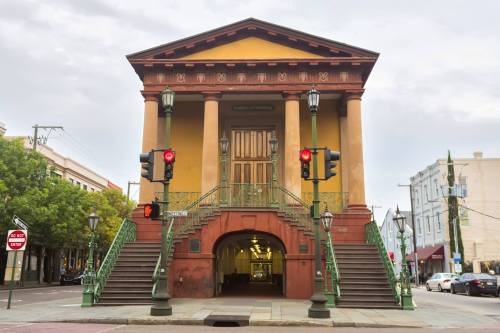 Historic building with a grand entrance, featuring columns and green railings, set against a cloudy sky. Traffic lights in view.