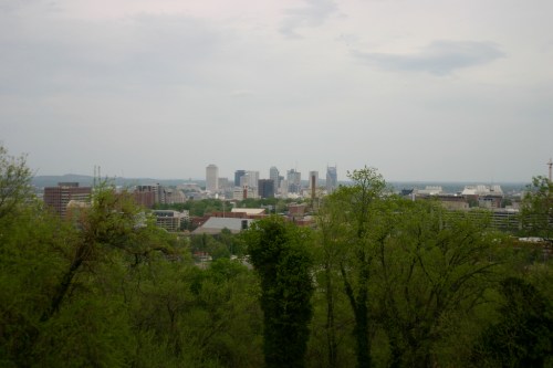 A city skyline is visible in the distance, surrounded by lush green trees under a cloudy sky.