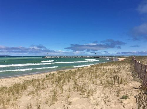 A sandy beach with green waves, a clear blue sky, and a distant pier under a few clouds.