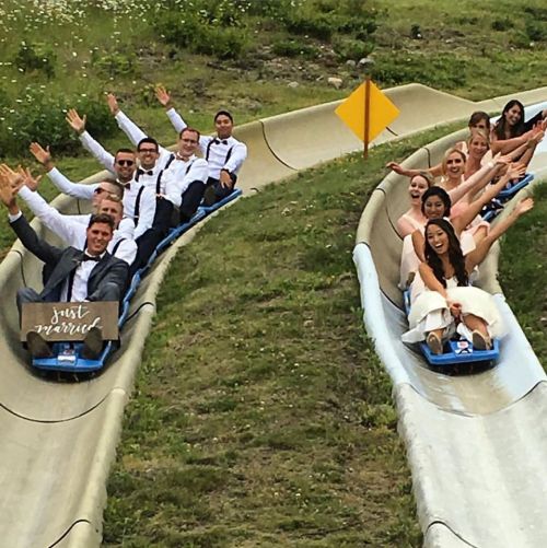 A group of people in formal attire joyfully riding a summer slide, waving and smiling as they descend.