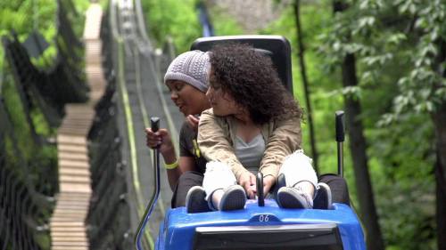 Two friends sit together on a ride, smiling, with a forested area and a bridge in the background.