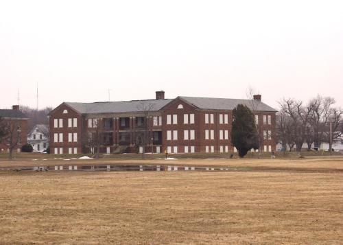 A large, brick building with multiple windows, set in a grassy area with a pond in the foreground and trees in the background.