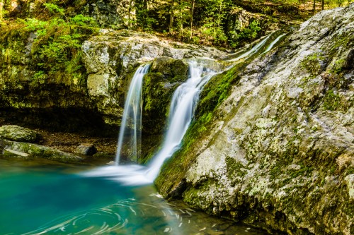 A serene waterfall cascades over moss-covered rocks into a tranquil turquoise pool, surrounded by lush greenery.