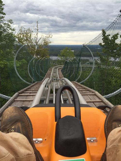 View from a roller coaster cart, showing the track winding down a hill surrounded by trees and a cloudy sky.