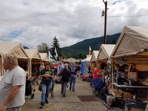 A bustling outdoor market with tents, shoppers, and mountains in the background under a cloudy sky.