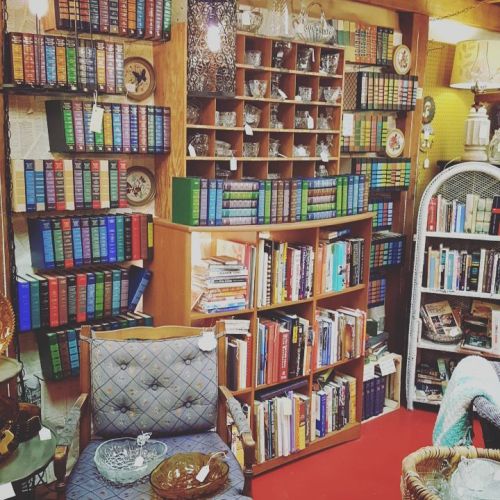 A cozy vintage bookstore with shelves of colorful books, glassware, and a comfortable chair in a warm, inviting space.