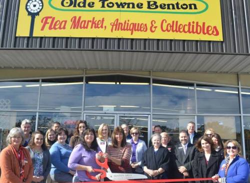A group of people gathered outside a flea market, holding a large red ribbon and scissors for a grand opening ceremony.