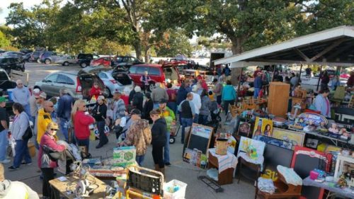A bustling outdoor flea market with many people browsing various stalls and items for sale under a shaded area.