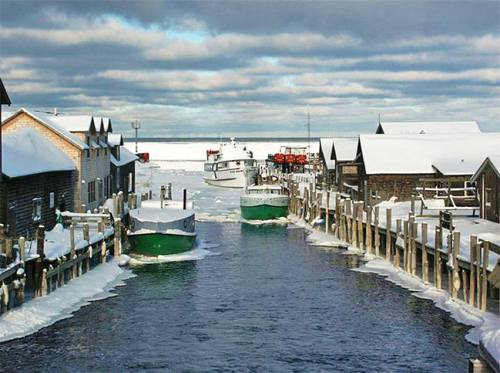 Snow-covered boats and wooden buildings line a frozen canal under a cloudy sky.
