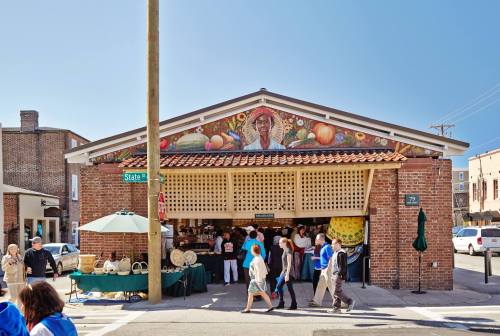 A vibrant market with a mural, featuring people walking by and stalls displaying fruits and crafts.